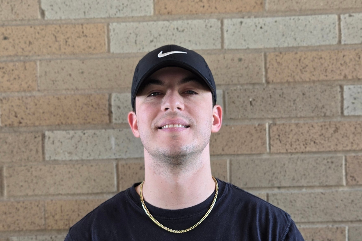 Jordan Schilling is pictured against a brick background, wearing a black Nike baseball cap, a gold chain, and a black t-shirt.